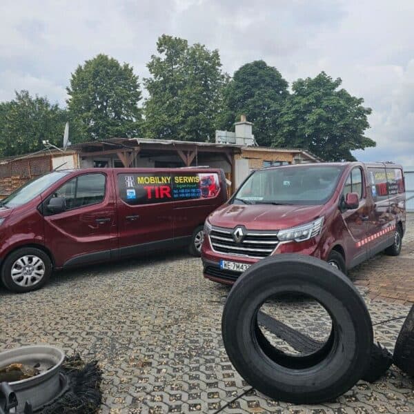 Dwa firmowe samochody osobowe zaparkowane obok siebie, stojące obok warsztatu. Obok widać także opony ciężarówki.