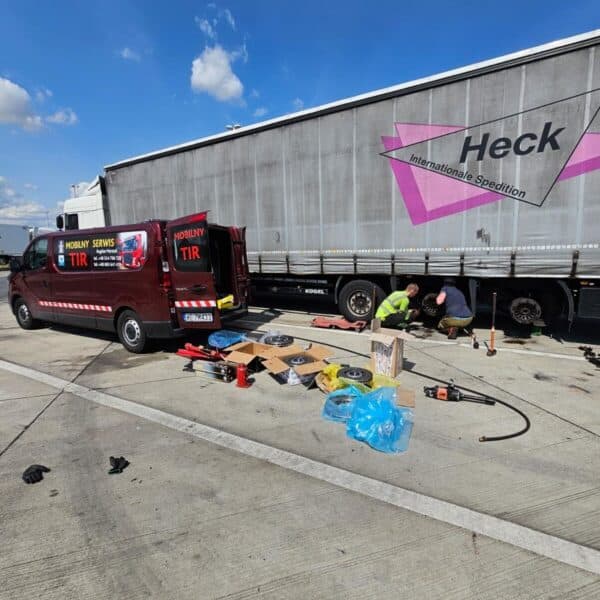 Dwóch mężczyzn naprawiających ciężarówkę. Obok stoi firmowy samochód.