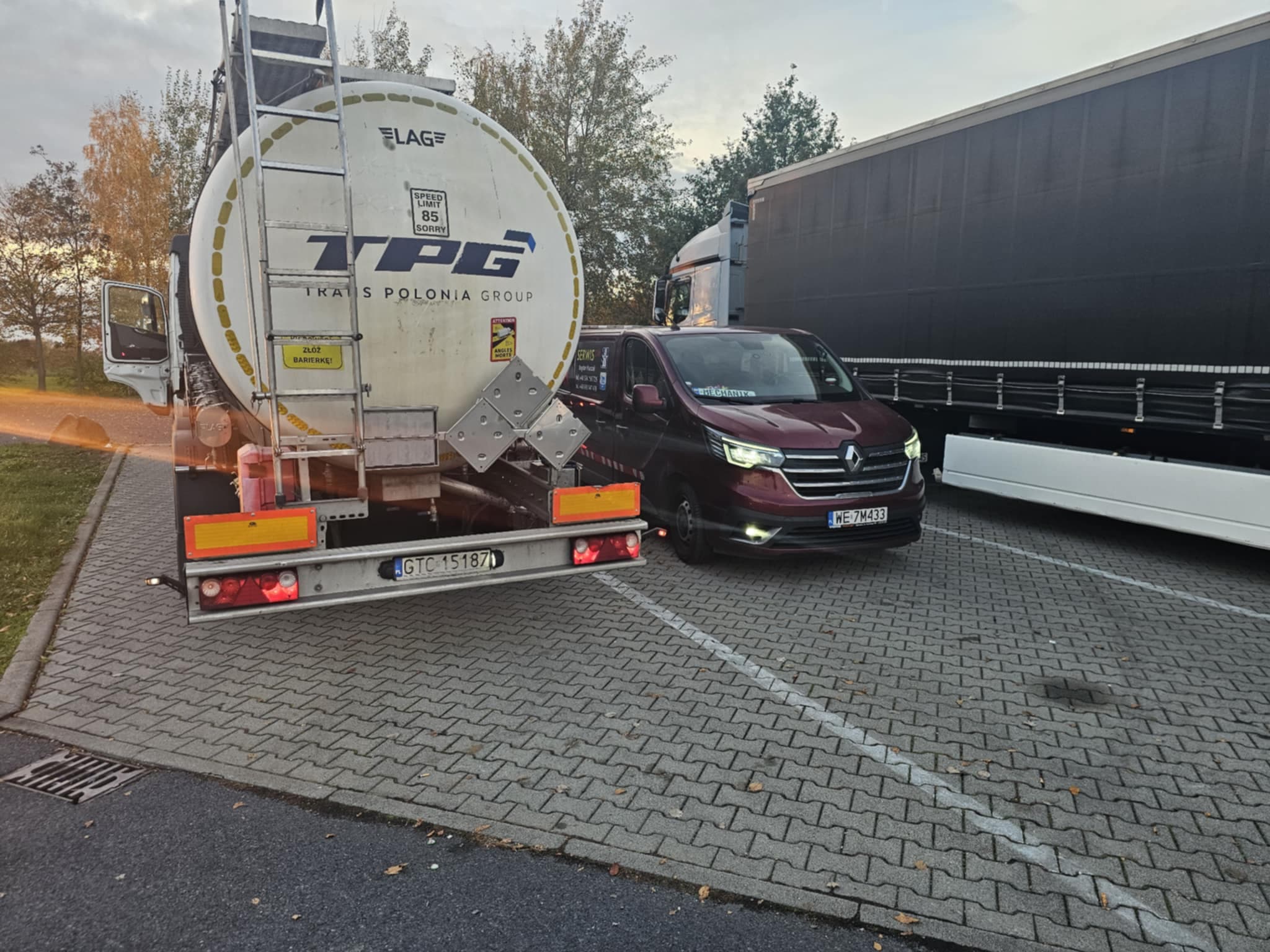 Samochód firmowy stojący obok ciężarówki z cysterną. Po prawej stronie stoi inna ciężarówka.